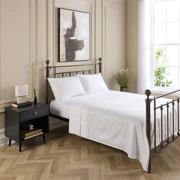 A metal framed bed in a modern bedroom. It features a white fitted sheet and flat sheet, with white pillowcases on pillows. There is a contemporary painting above the bed and a small dark wood cabinet next to the bed. The floor is a wooden geometric pattern.