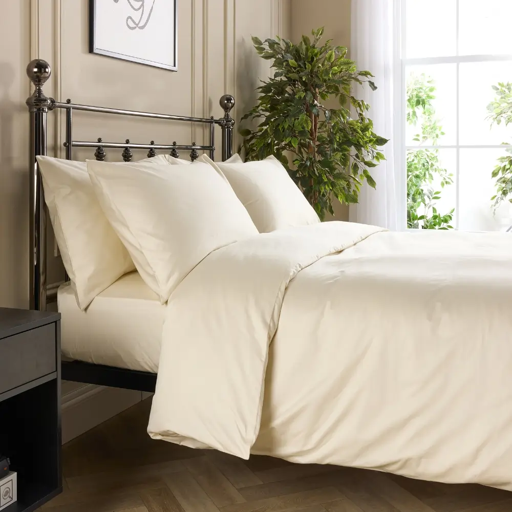 A side view of a hotel quality cream coloured cotton duvet cover and pillowcases covering a duvet and pillows on a metal framed bed in a contemporary bedroom.