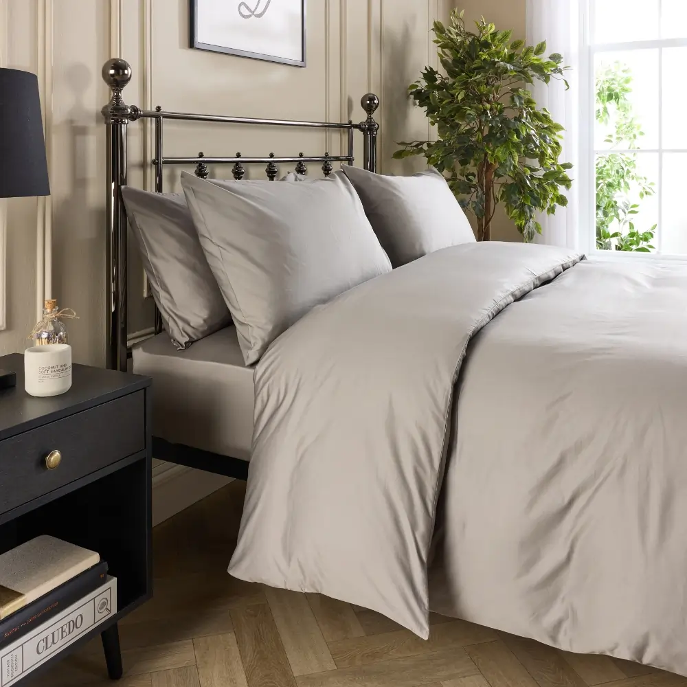 A side view of a hotel quality grey coloured cotton duvet cover and pillowcases covering a duvet and pillows on a metal framed bed in a contemporary bedroom.