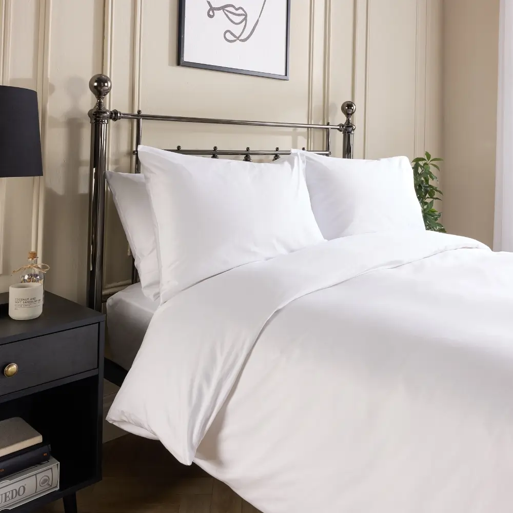 A side view of a hotel quality white cotton duvet cover and pillowcases covering a duvet and pillows on a metal framed bed in a contemporary bedroom.