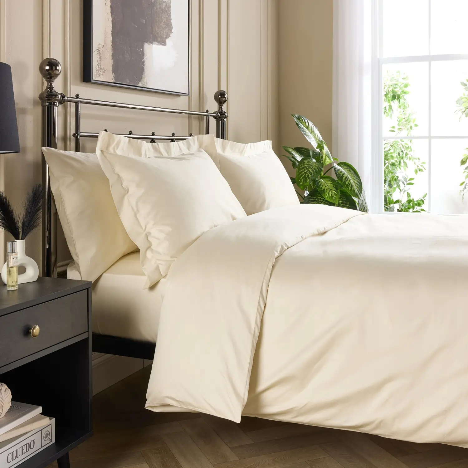 A side view of a hotel quality cream coloured cotton 480 thread count duvet cover and pillowcases covering a duvet and pillows on a metal framed bed in a contemporary bedroom.