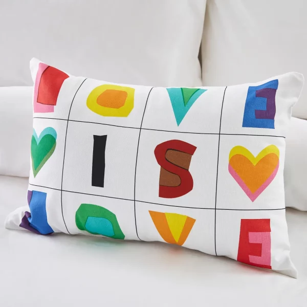 A white cushion from Martex Pride. It features a series of squares with multi-coloured letters and love hearts in them, spelling out the slogan 'Love is Love'. It's sitting on a bed.