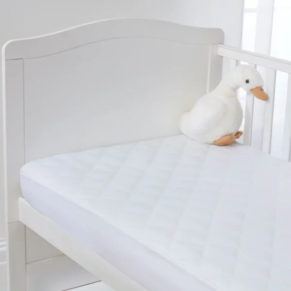 Clair De Lune anti-allergy quilted mattress protector on a mattress on a cot bed. There is a white plush duck toy with an orange beak and feet sitting on the mattress near the right side of the crib. The crib has a curbed headboard and slatted sides.