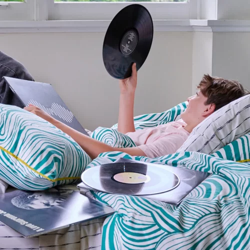 A teenage boy relaxing on a green zebra marble teal duvet cover. He is holding up a vinyl record and smiling at it.