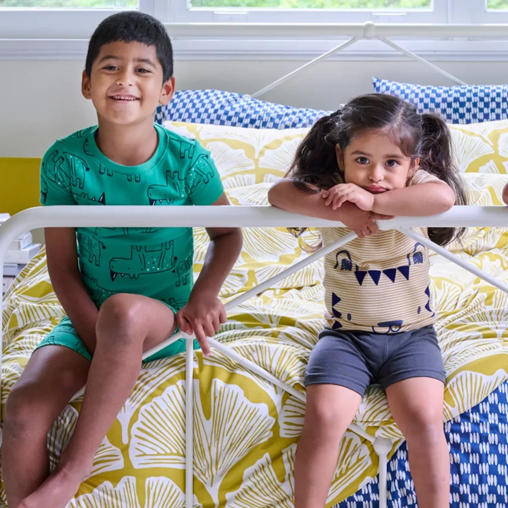 Two kids sitting on the end of a bed that has a golden mustard feather palm patterned duvet cover on it.