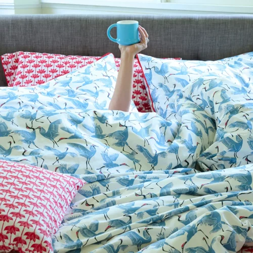 A blue patterned family of cranes duvet cover and pillowcase set. There is a hand coming up out of the pile of bedding that's holding a blue coffee cup.