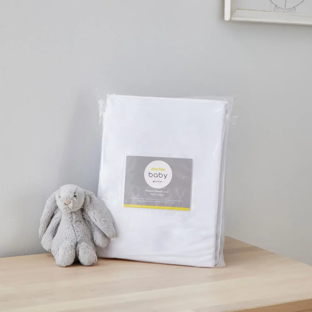 A white moses basket fitted sheet from Martex Baby in its packaging. There is a grey toy bunny next to it.