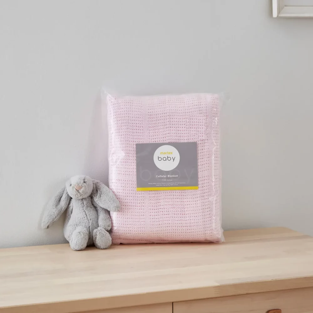 A pink cellular blanket from Martex Baby in its packaging. There is a grey toy bunny sitting next to it.