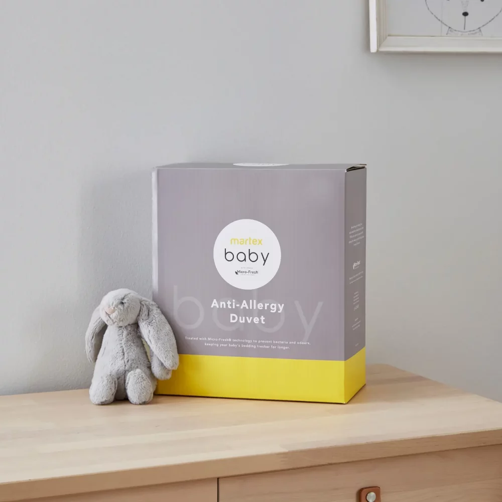 Grey and yellow packaging for a Martex Baby anti-allergy 4 tog duvet for use in a cot. The packaging is sitting on a chest of drawers and there is a small grey toy bunny sitting next to it.