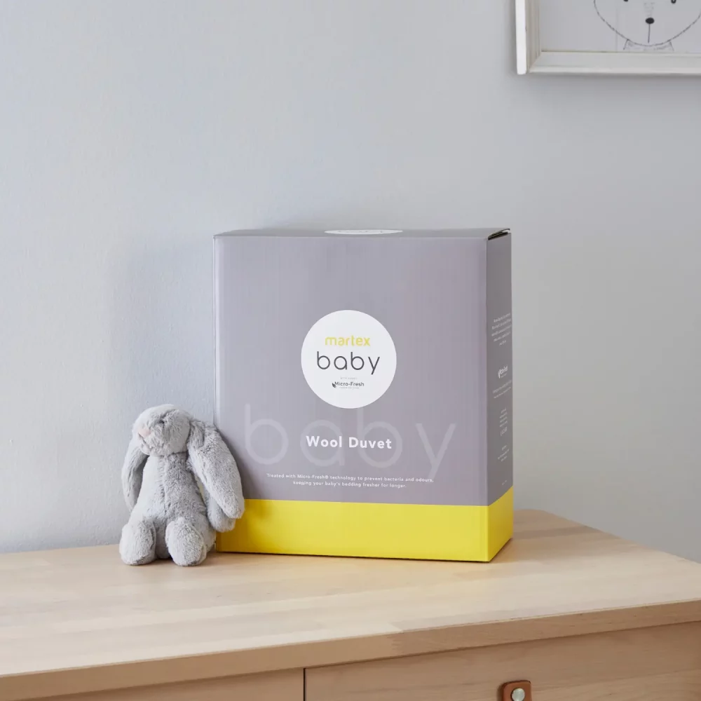 Grey and yellow packaging for a Martex Baby 4 tog wool duvet for use in a cot bed. The packaging is sitting on a chest of drawers and there is a small grey toy bunny sitting next to it.