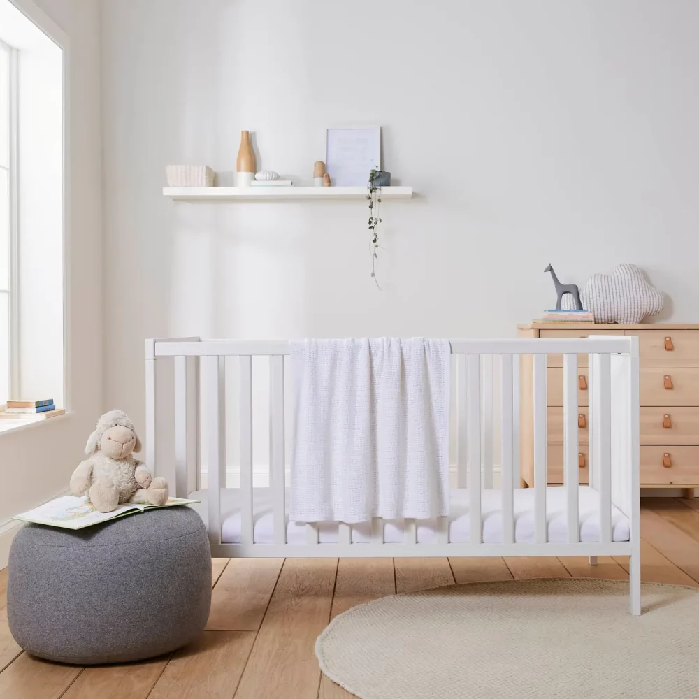 A white cellular blanket hanging off the edge of a a cot in a child's bedroom. The blanket is part of the Martex Baby cot bed starter set, the rest of which is not pictured.