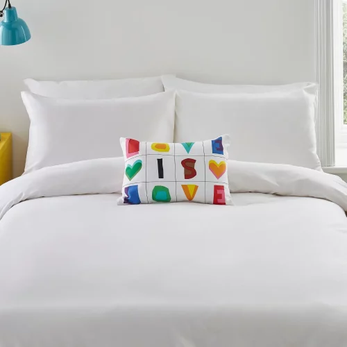 A white cushion from Martex Pride. It features a series of squares with multi-coloured letters and love hearts in them, spelling out the slogan 'Love is Love'. It's sitting on a bed.