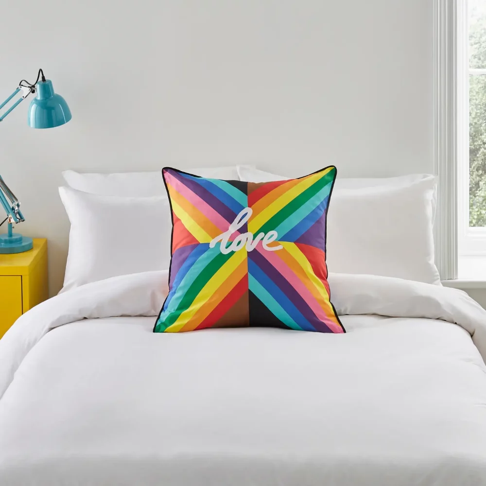 A multi-coloured rainbow cushion from Martex Pride. It has the word 'Love' in the middle of it. It's sitting on a bed.